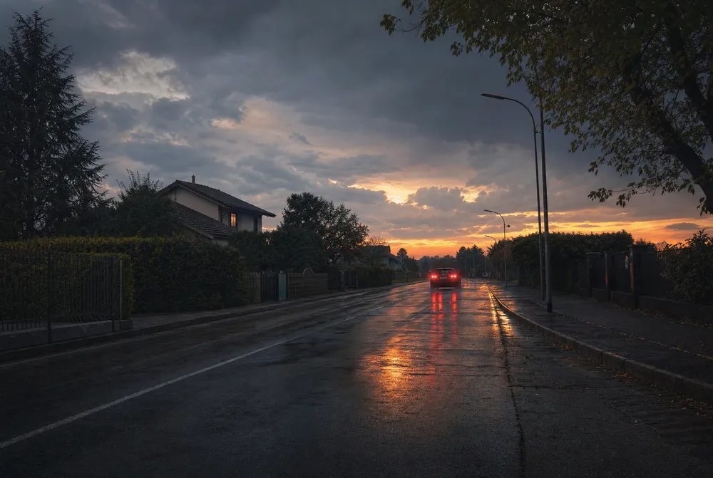 A wet suburban road at dusk with tail lights reflecting on the asphalt